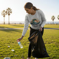 A woman in a park on a sunny day, wearing a light gray sweatshirt with a wave graphic and the words 'WAVE SPACE CAN', black pants, and holding a black trash bag while picking up a plastic water bottle from the grass.